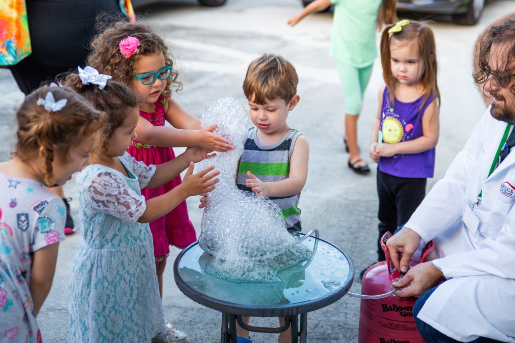Group of children touching a large cluster of helium bubbles on a table outside.
