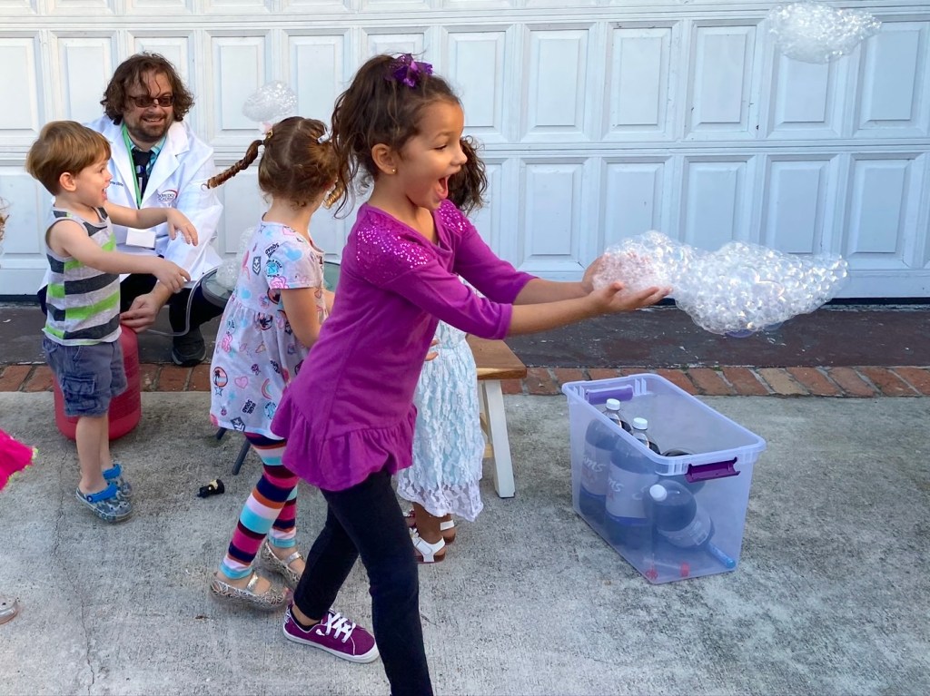 Children laughing and reaching for helium bubble clusters during an outdoor science activity.