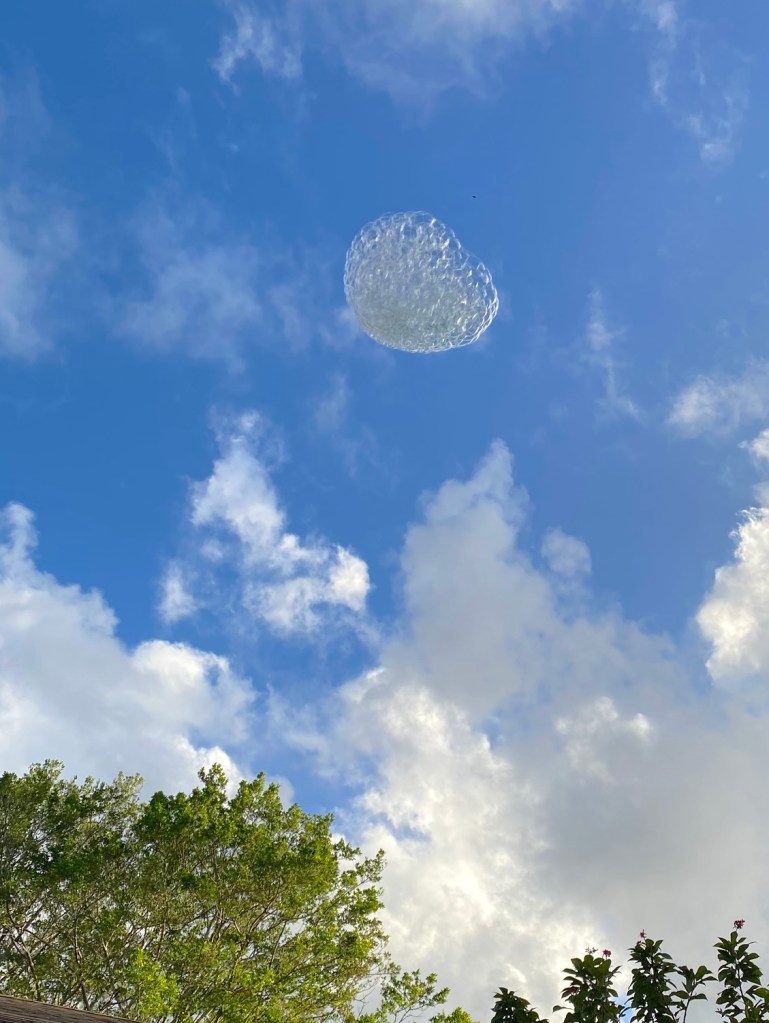 Large helium bubble cluster floating high against a blue sky with clouds.
