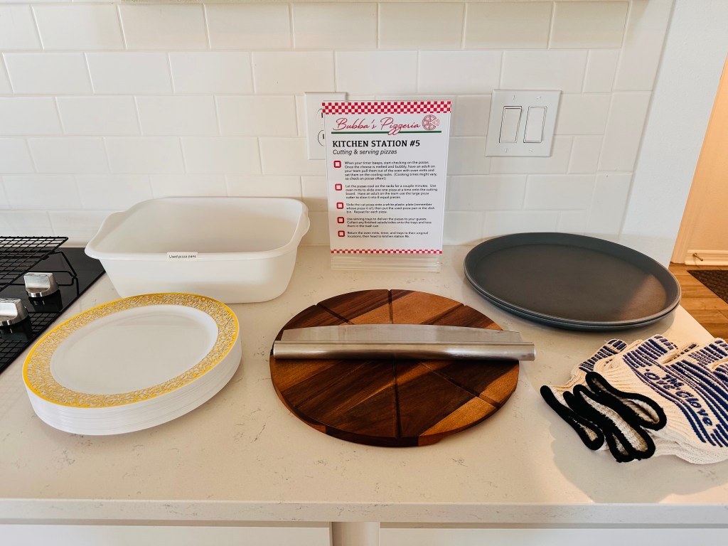 Kitchen Station #5 setup with plates, pizza cutting board, cutter, oven mitts, and instruction sheet for serving pizzas.