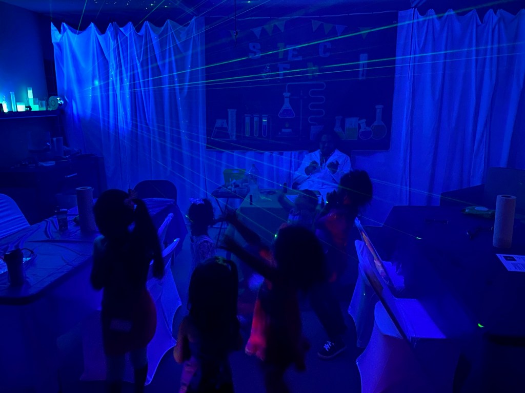 Children move through a darkened garage filled with green laser beams from a star projector, with blacklight-lit science decor behind them.