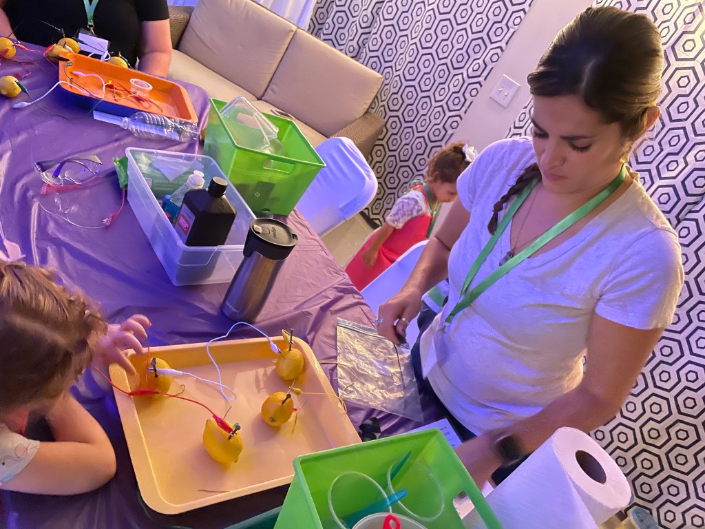 A woman helps children connect wires to lemons to create a lemon battery, with trays of lemons and science supplies on a purple-covered table.