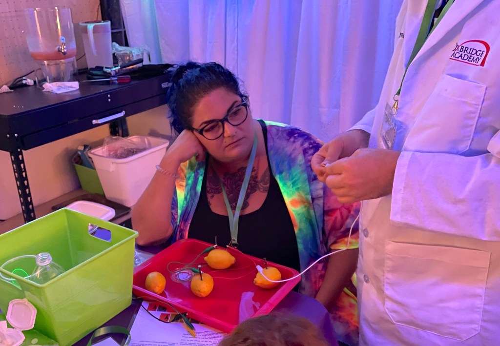 A woman in a tie-dye jacket watches as a person in a white lab coat demonstrates connecting wires to lemons for a lemon battery experiment.