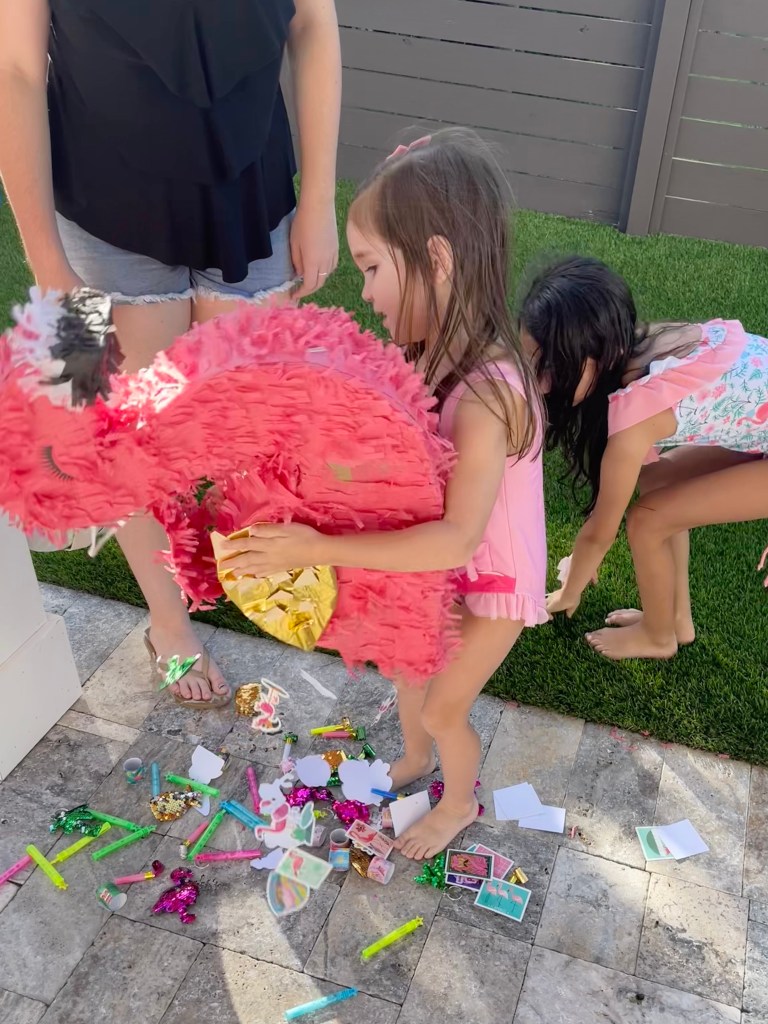 Young girl pulls toys from a broken flamingo piñata while another child gathers prizes from the ground.