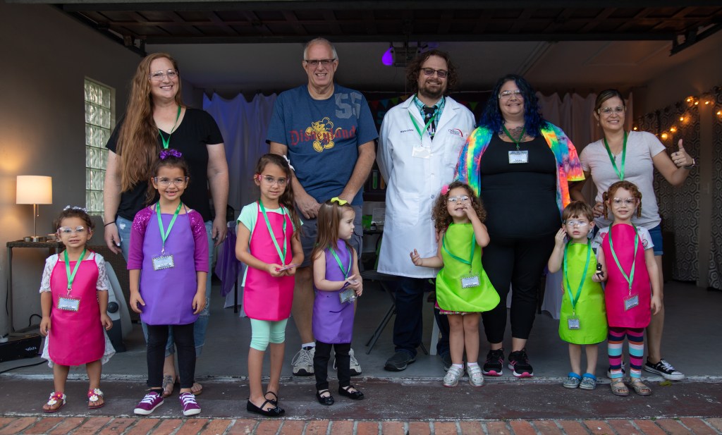 Group photo of seven young children wearing safety goggles, colorful aprons, and science-themed name badges, standing in front of four adult helpers and Wayne the rocket scientist in a white lab coat.