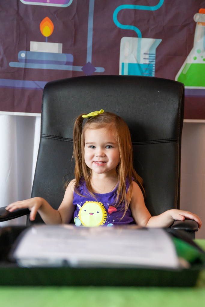 Toddler girl in a purple space-themed tank top sitting in a black chair in front of a science-themed backdrop with colorful beakers and flasks.