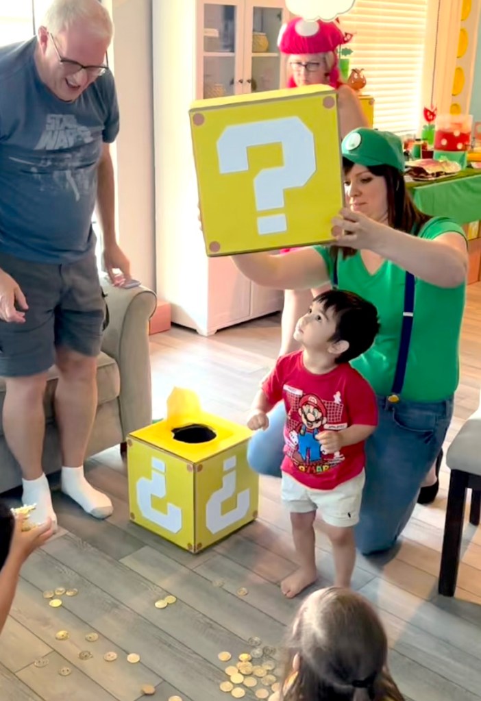 A woman dressed as Luigi holds a large yellow Mario question block above a young boy’s head while gold coins spill onto the floor during a Super Mario-themed birthday party.