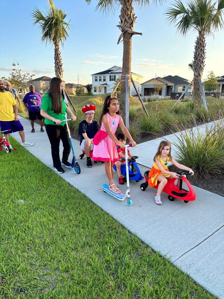 Kids and adults dressed as Mario Kart characters racing scooters and ride-on toys down a neighborhood sidewalk lined with palm trees.