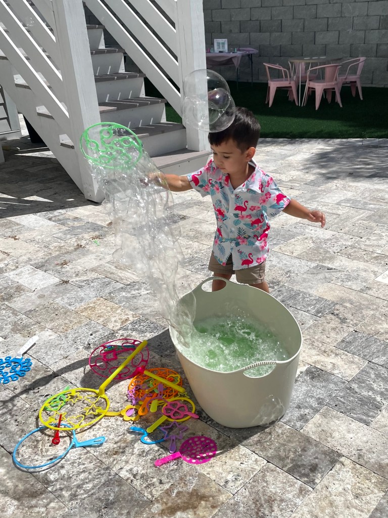 Toddler boy wearing a flamingo shirt plays with oversized bubble wands at an outdoor station.