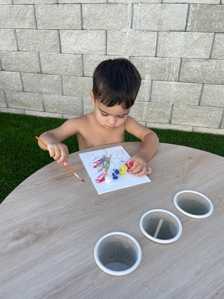 A young boy sits at a small round table, focused as he paints colorful abstract lines on a canvas with paint pots nearby.
