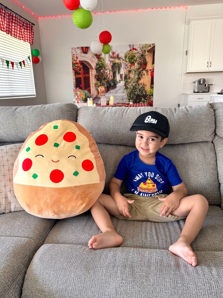 Noah sitting on a gray couch at his Bubba’s Pizzeria birthday party, smiling in a “Any Way You Slice It I’m Awesome” pizza shirt and black cap, next to a giant plush pizza pillow with red and green toppings.