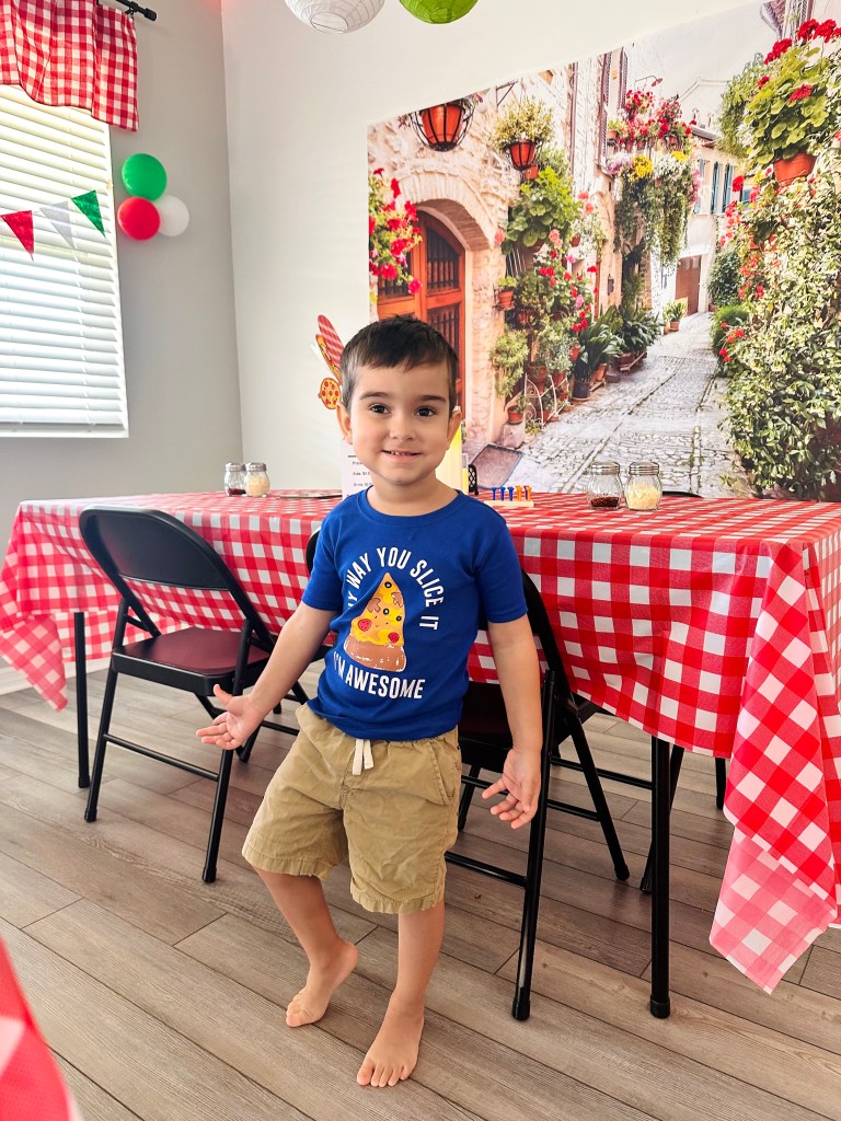 A young boy smiling at the camera while standing barefoot in front of a table with a red-and-white checkered tablecloth, Italian-inspired wall mural, and pizza-themed party decorations.