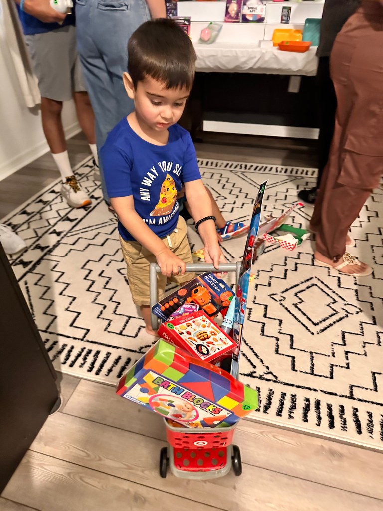 A little boy in a blue pizza-themed shirt pushes a red toy shopping cart filled with prizes, including blocks, a dart blaster, and art kits.