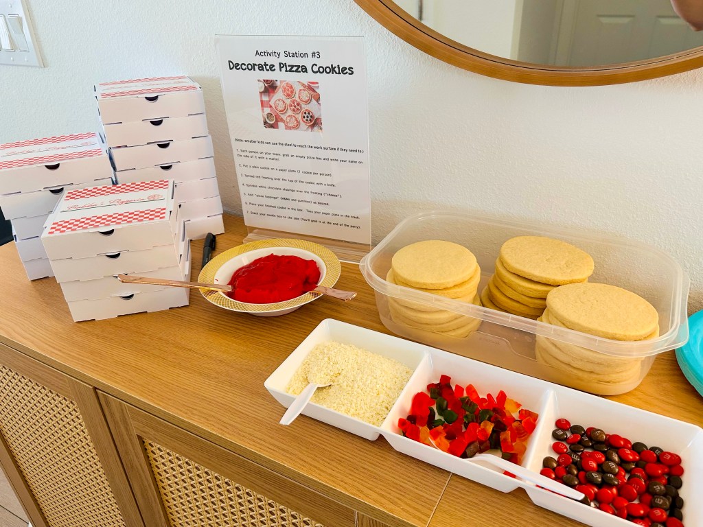 A pizza cookie decorating station with stacked mini pizza boxes, plain sugar cookies, red frosting, white chocolate shavings, gummy candy, and M&Ms.