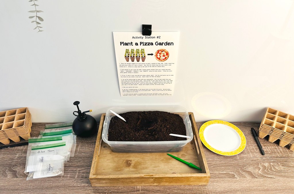 Overhead view of the Plant a Pizza Garden station, showing a bin of soil, seed packets, planting trays, and a sign with instructions.
