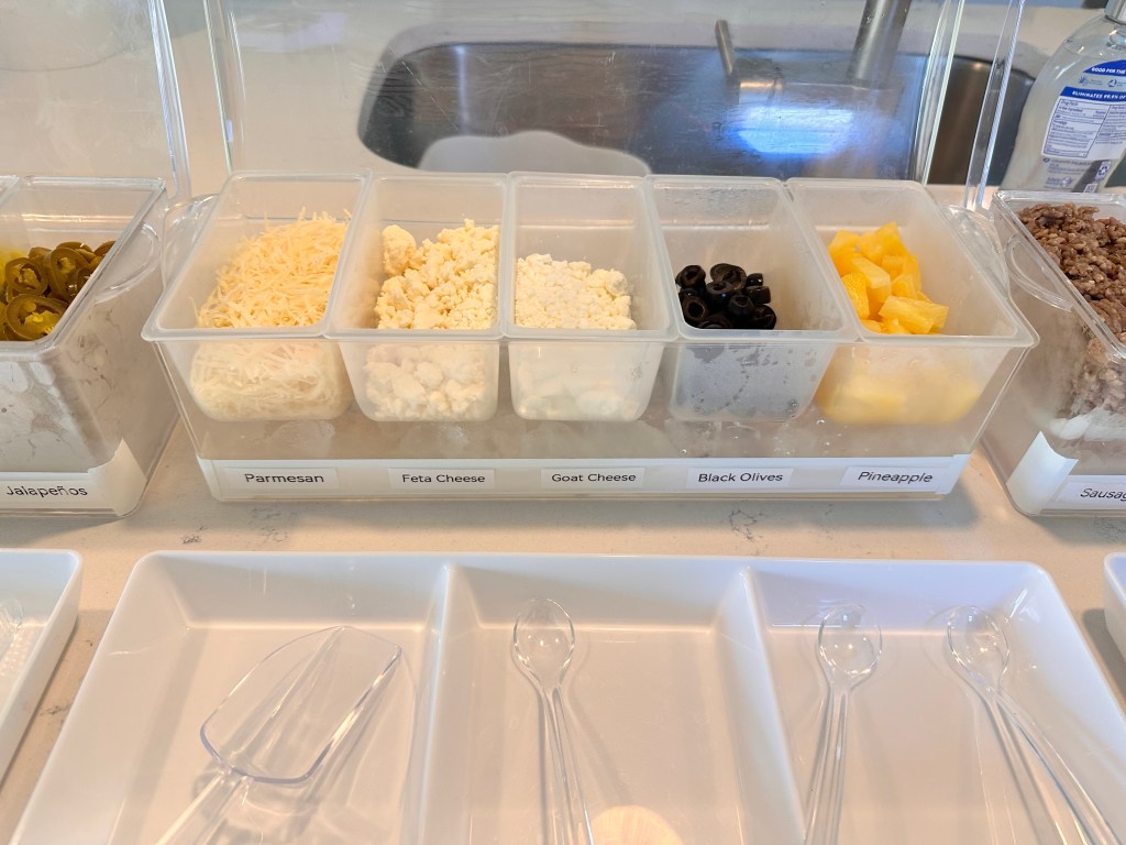 Pizza topping containers filled with parmesan, feta cheese, goat cheese, black olives, and pineapple.