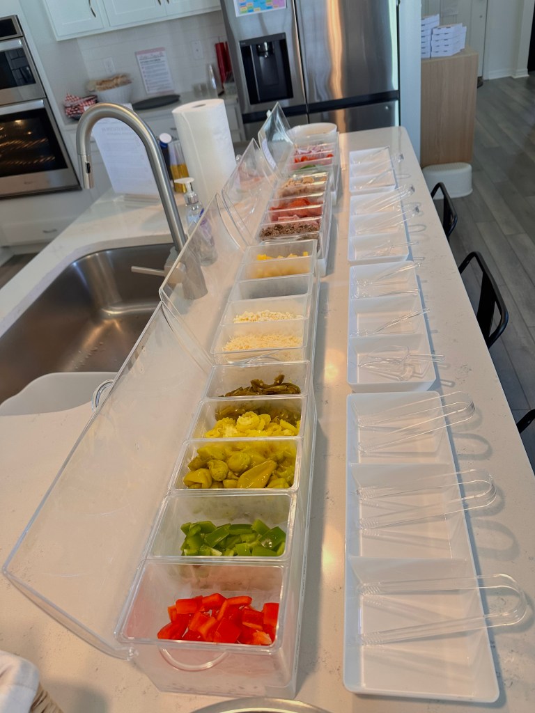 Clear bins filled with colorful pizza toppings such as cheese, peppers, and meats lined up on a kitchen counter with serving utensils.