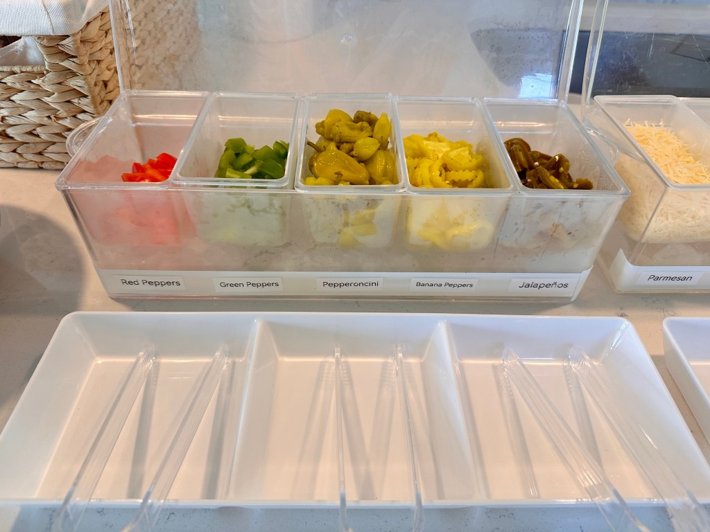 Close-up of pizza topping containers with red peppers, green peppers, pepperoncini, banana peppers, and jalapeños.
