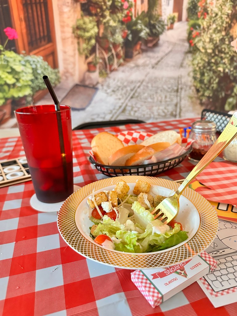 Salad with lettuce, tomatoes, feta cheese, and croutons served on a red checkered tablecloth with bread basket and drink