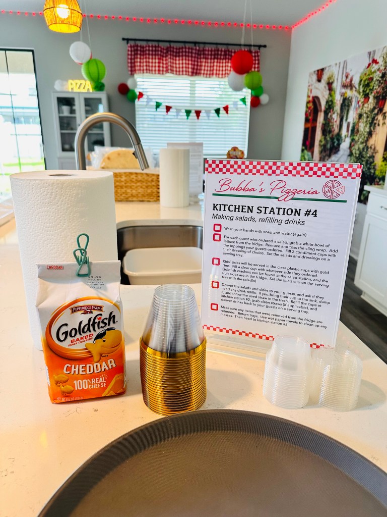 Party kitchen station with Goldfish crackers, clear plastic cups, paper towels, and printed salad-making instructions for a pizza-themed birthday.