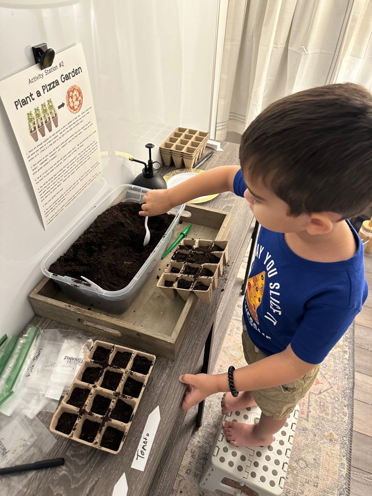A young boy stands on a stool scooping soil into seed starter trays at the Plant a Pizza Garden activity station, which features seed packets, a watering mister, and an instructional sign.