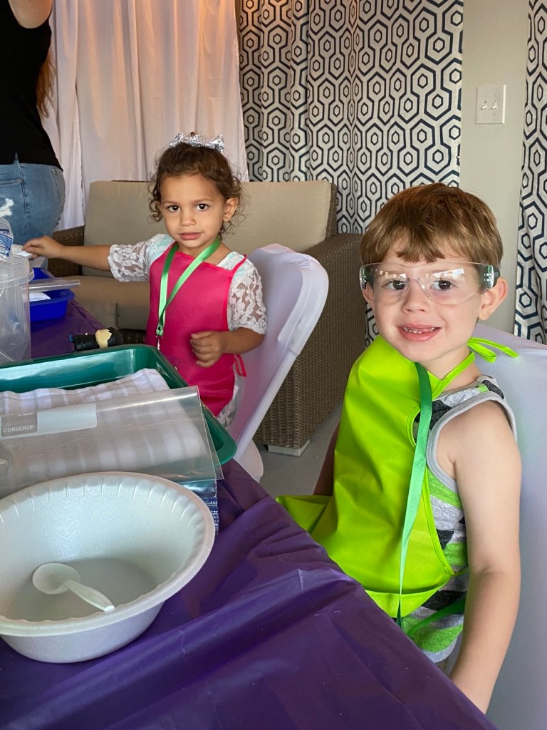 A young boy and girl, both wearing safety goggles and aprons, sit at a table with trays and supplies, smiling as they prepare for the ice cream-making activity.