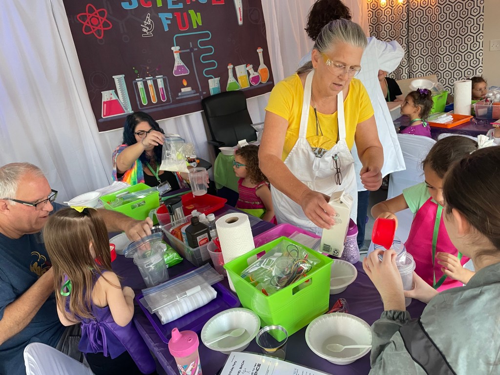 Mallory’s grandma wearing a yellow shirt and white apron pours heavy cream while helping children make ice cream during a science-themed party activity.