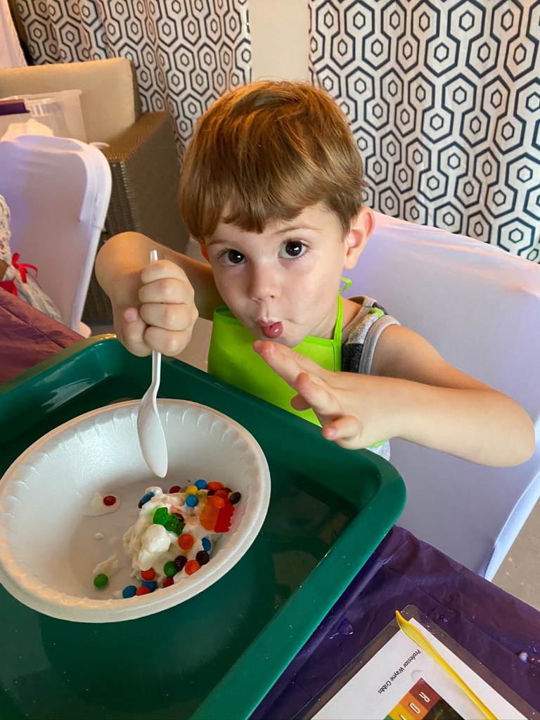 Young boy in a green apron enjoys homemade ice cream topped with colorful candy and gummy bears during the party’s science experiment.