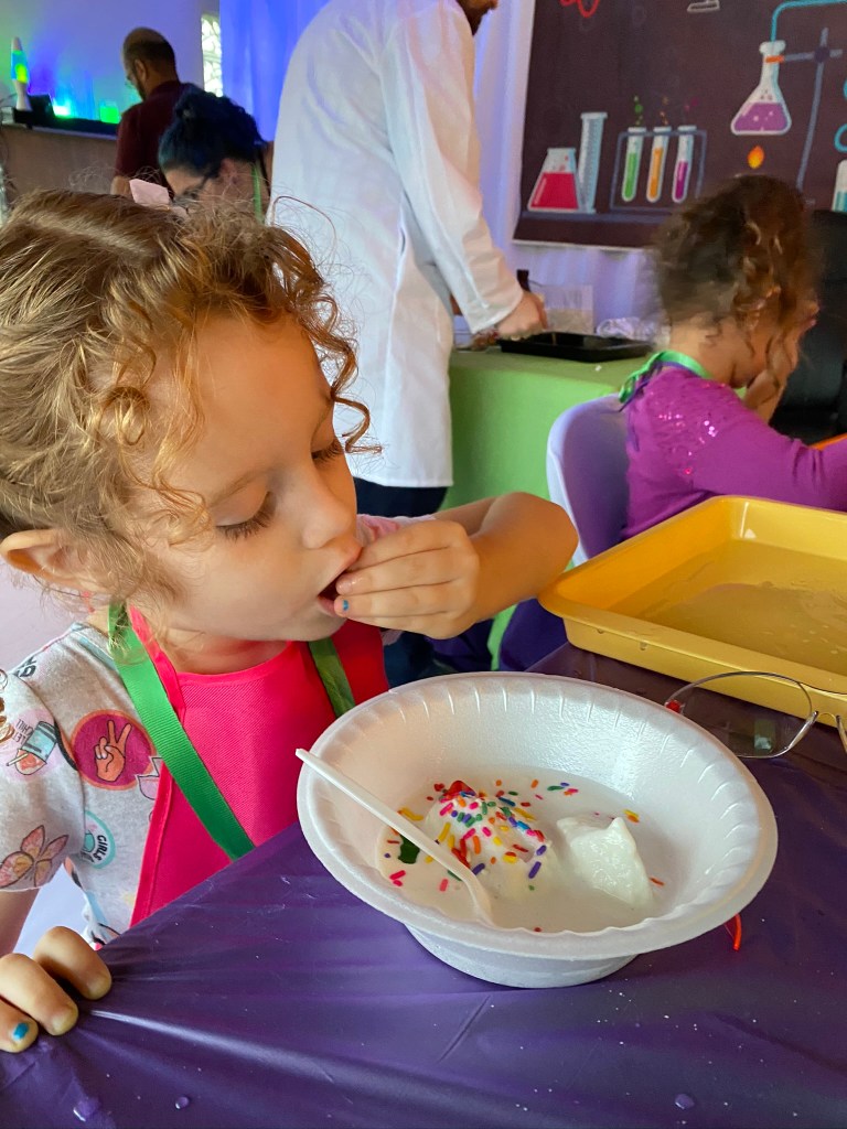 Young girl with curly hair and a pink apron eats homemade ice cream topped with rainbow sprinkles at the science party.
