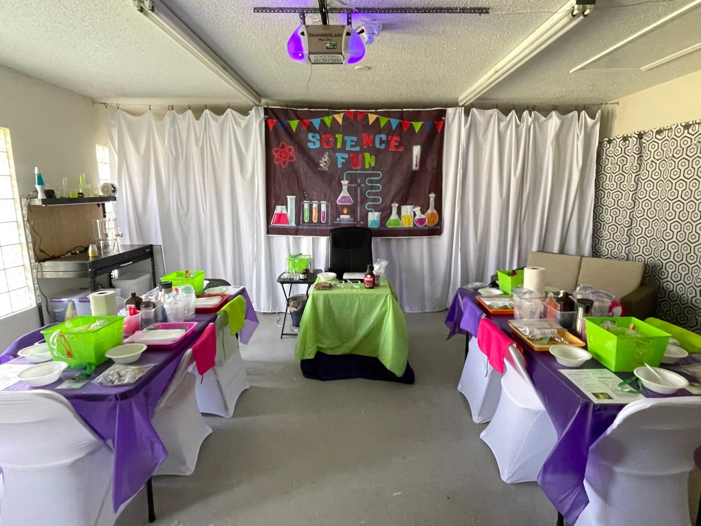 Science-themed birthday party setup in a decorated garage with tables, chairs, and banner.