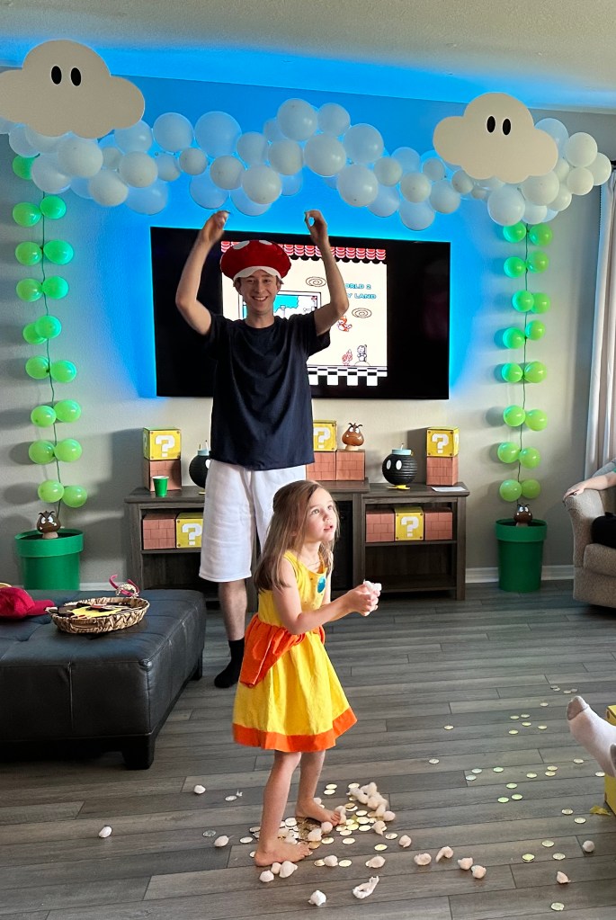 A child in a yellow dress stands in a pile of gold coins and cotton balls that spilled from a Super Mario punch block.