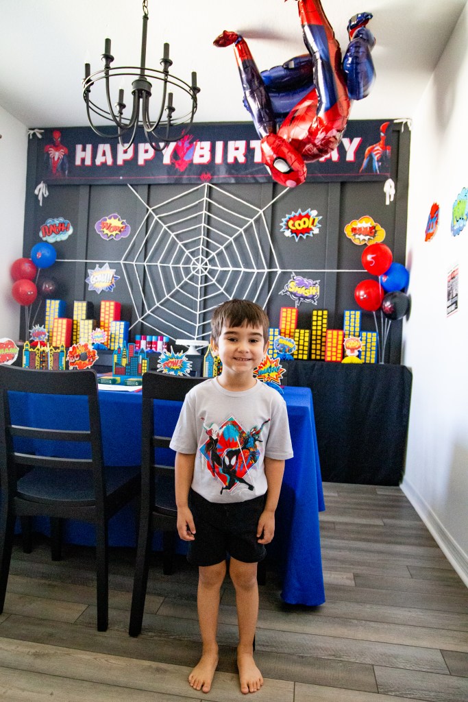 Young boy smiling in front of a Spider-Man birthday party backdrop with a giant web, city skyline decorations, and a hanging Spider-Man balloon