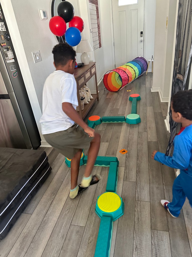 Kids running across a balance beam setup leading into a rainbow tunnel in hallway during Spider-Man obstacle course.