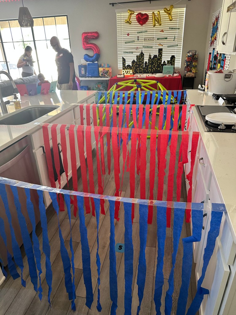 Spider-Man party obstacle course station with red and blue streamers taped across kitchen counters as a crawl-through tunnel.