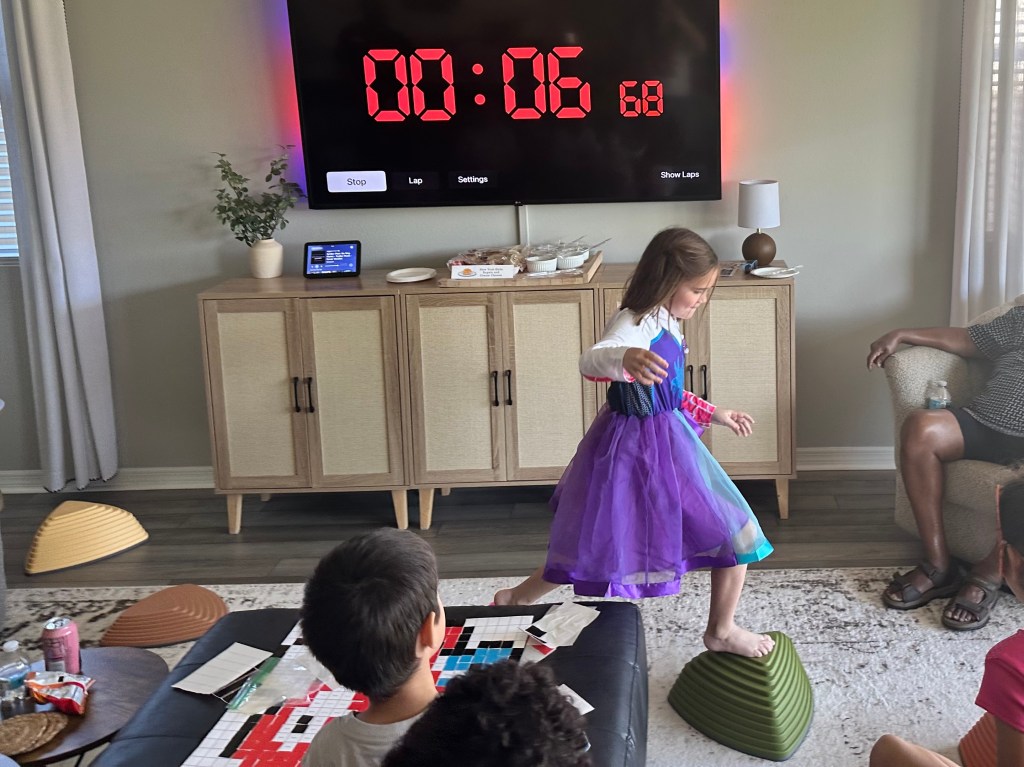 Child balancing on stepping stones during Spider-Man party obstacle course with large timer on TV in background.