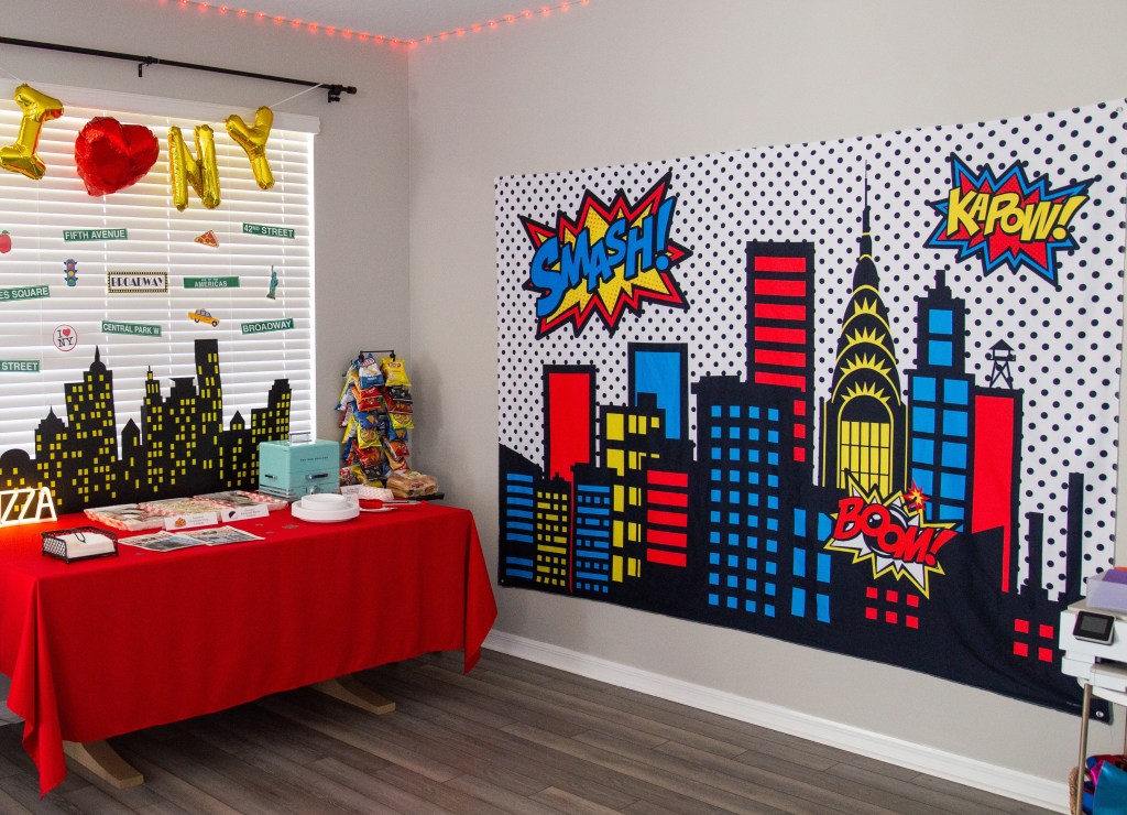 Oversized comic book city backdrop with “Smash!” and “Kapow!” speech bubbles set up next to a Spider-Man party food table.