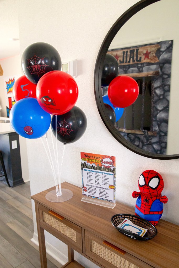 Table setup with Spider-Man balloons, a superhero name chart, a plush Spider-Man doll, and a basket of name tag stickers at a birthday party entrance.