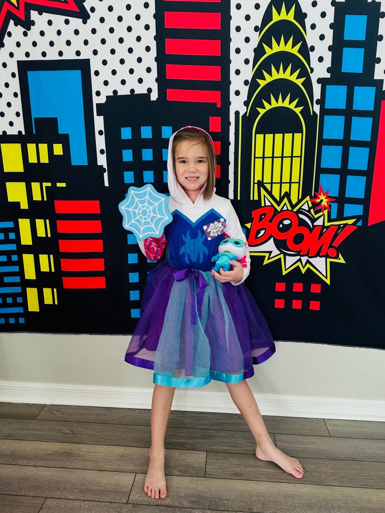 Child dressed as Spider-Gwen posing in front of a bright comic book skyline backdrop at a Spider-Man birthday party.