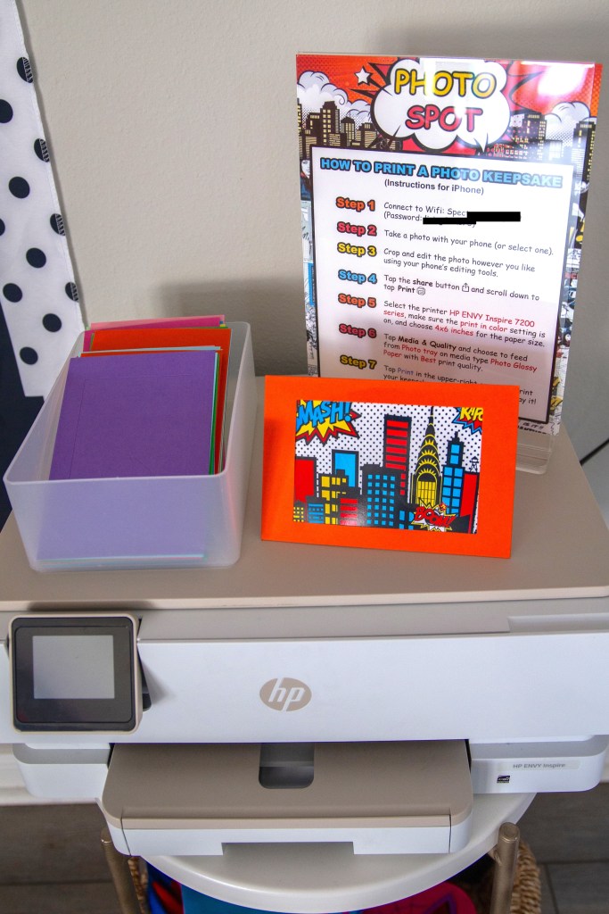 Printer station with colorful cardstock frames, photo booth instructions, and comic-style decor for a Spider-Man birthday party.