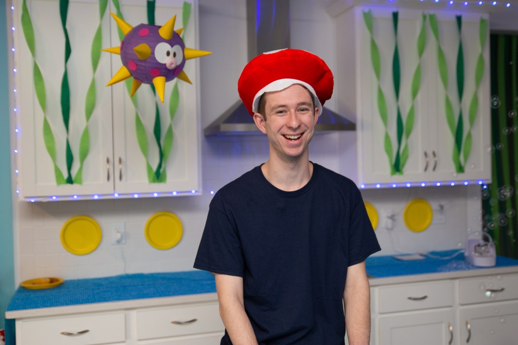 Smiling boy wearing a Toad hat standing in front of Water Land decorations.