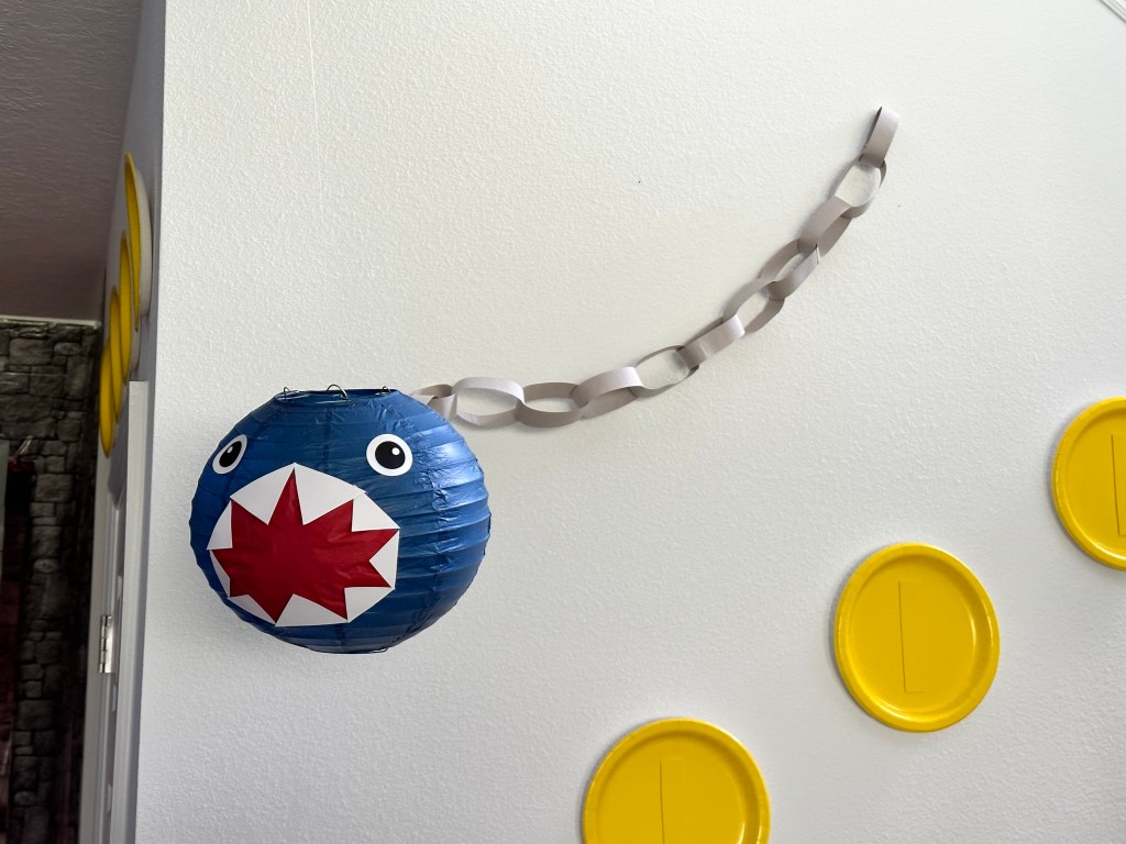 DIY Chain Chomp decoration made from a blue paper lantern with white teeth, red mouth, and paper chain links for the chain, hanging on the wall with paper plate Mario coins nearby.