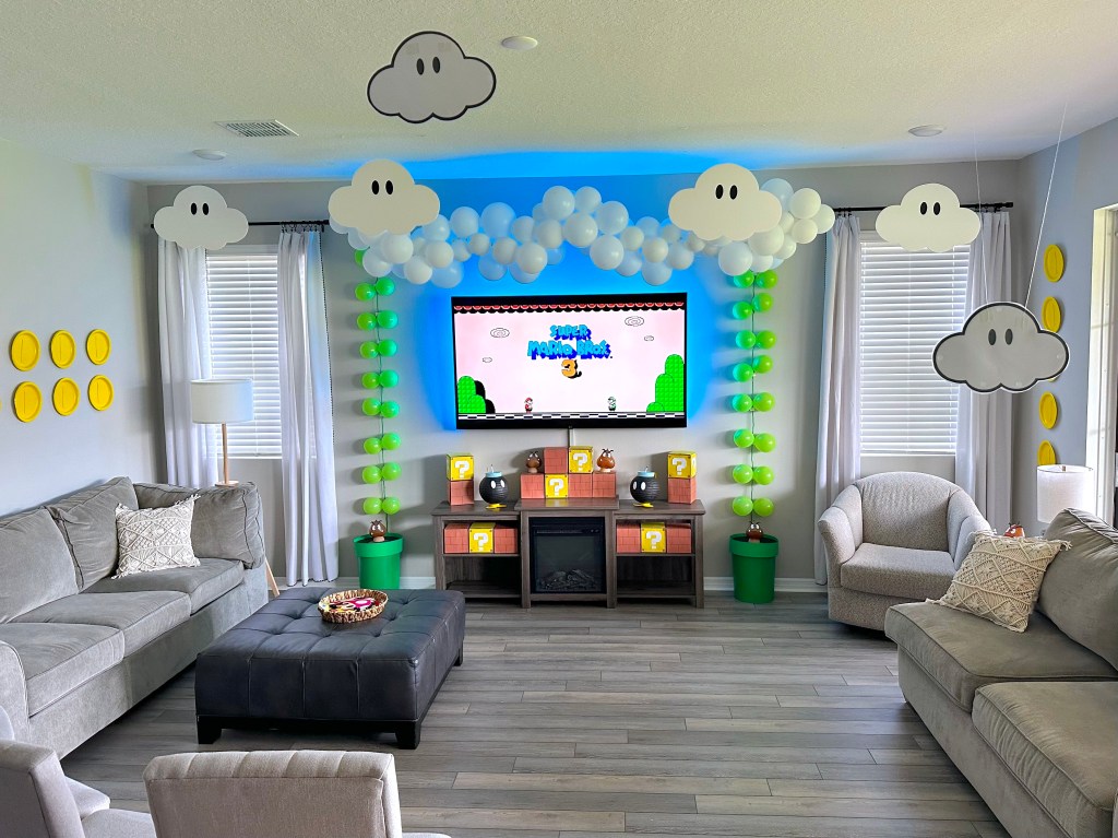 Wide shot of the living room transformed into Sky Land from Super Mario 3, with balloon vines, foam board clouds, hanging paper coins, and a TV displaying the game screen.