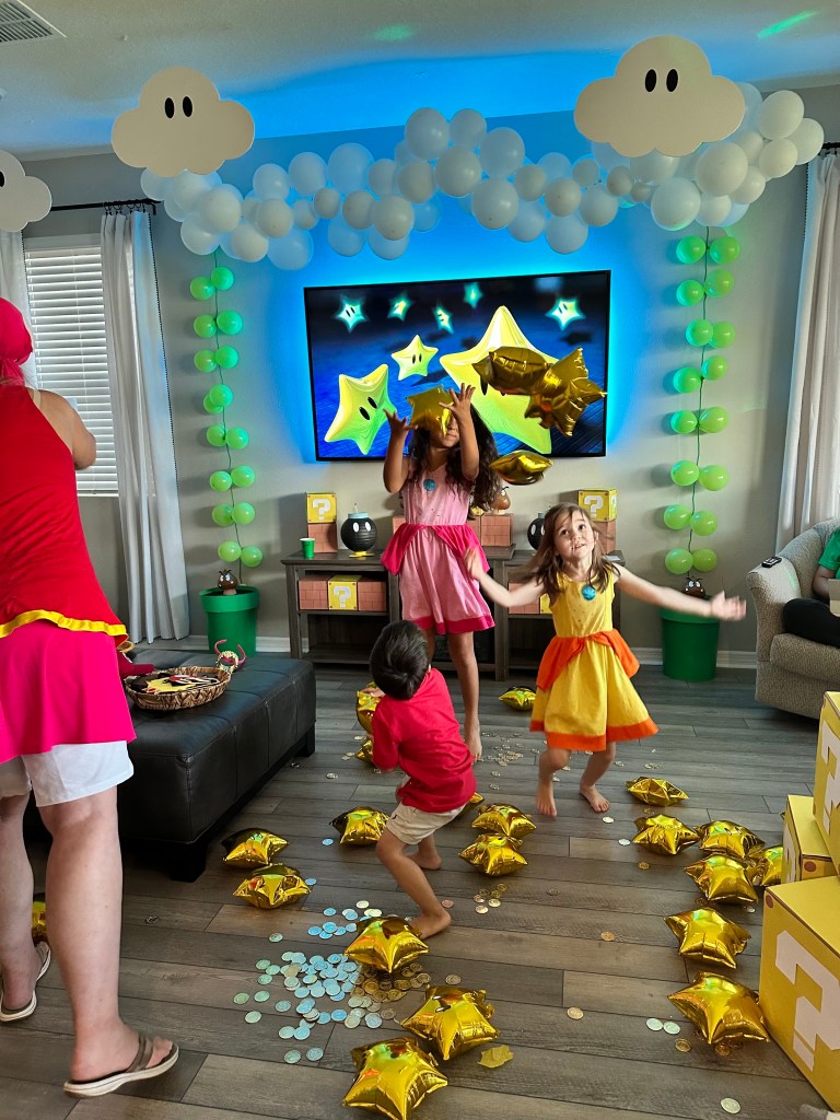 Three children dressed as Mario, Princess Peach, and Princess Daisy joyfully play in a living room decorated with Super Mario-themed props, tossing and dancing among gold star balloons and scattered gold and silver coins.