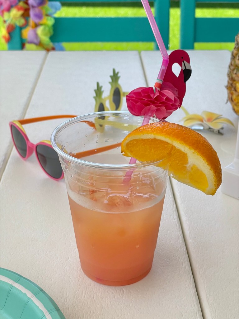 Cup of orange and pink layered tropical drink garnished with an orange slice and a flamingo paper straw.
