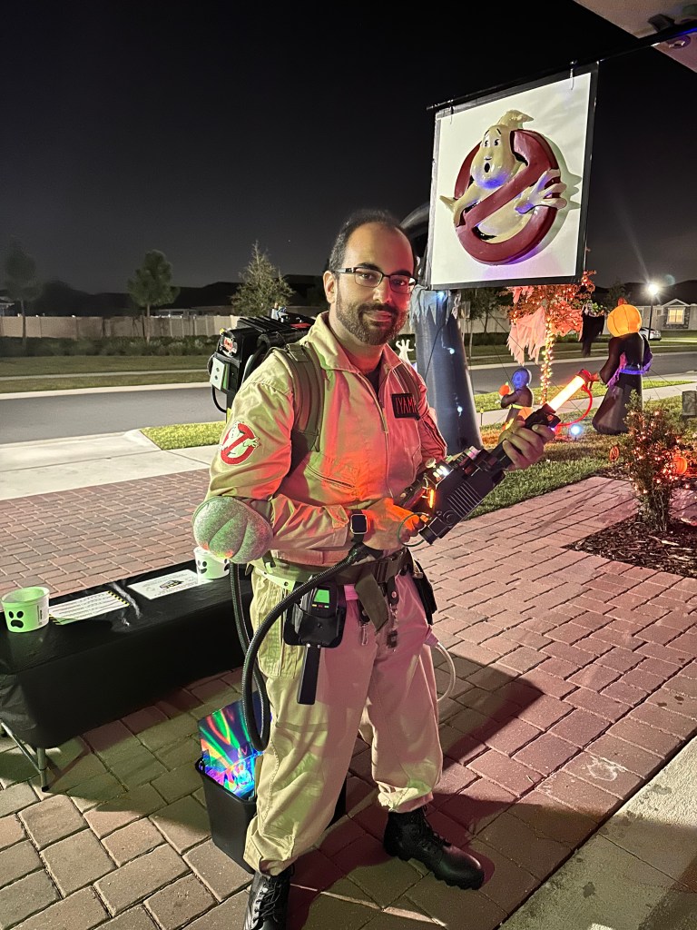 Man in full Ghostbusters costume holding a lit-up proton blaster at night in front of a themed Halloween garage display.