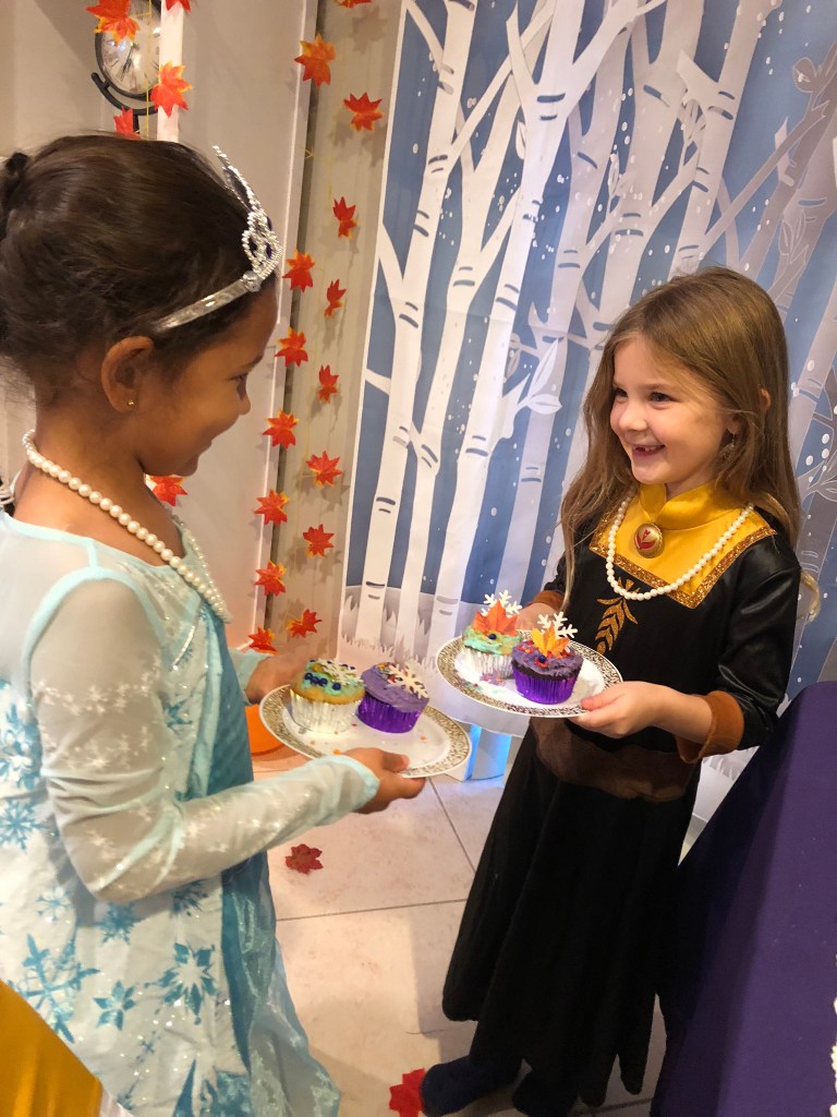 Two girls dressed as Anna and Elsa hold plates of decorated cupcakes, each featuring colorful frosting and themed toppers like snowflakes and fall leaves.