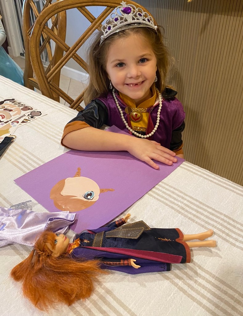 A smiling girl dressed as Anna sits at the craft table with her finished Anna sticker art and matching Anna doll.