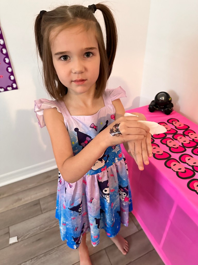 Young girl in a Kuromi dress carefully pressing a wet paper towel over a Sanrio tattoo on her hand at a pink tattoo station.