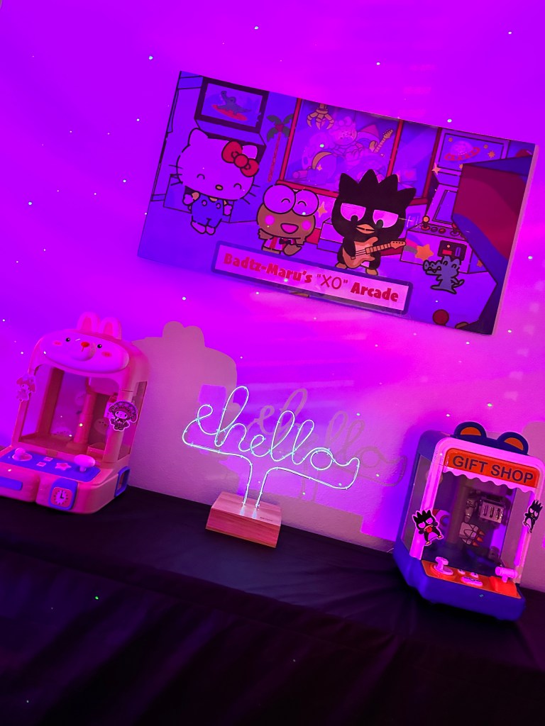 Badtz-Maru-themed arcade station with two mini claw machines, a glowing “hello” sign, and a custom poster with Hello Kitty characters under purple party lighting.