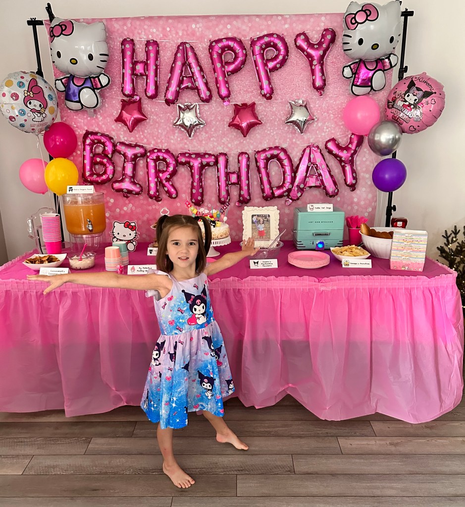 A young girl in a Kuromi and My Melody dress poses with arms outstretched in front of a bright pink Hello Kitty-themed birthday table, featuring a “Happy Birthday” balloon backdrop, snacks, cake, and drinks.